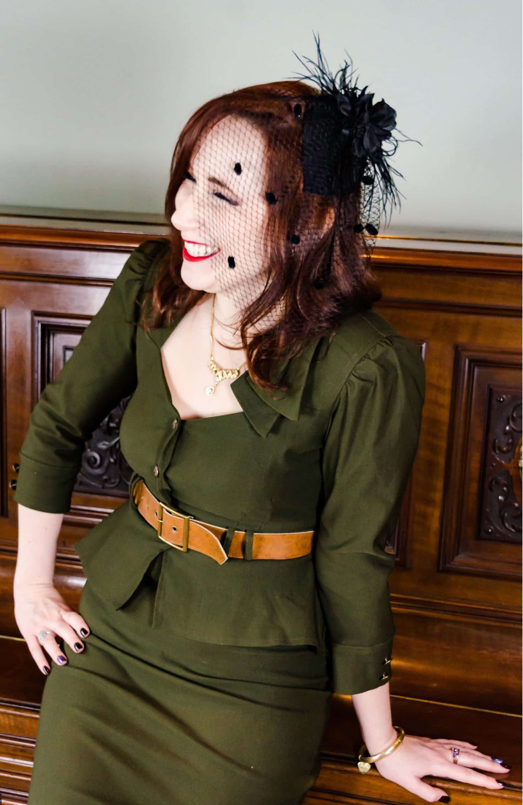 A laughing woman dressed in a vintage-style olive green peplum suit, a wide brown belt, and a black feathered fascinator with a birdcage veil, leaning against an ornate wooden piano.