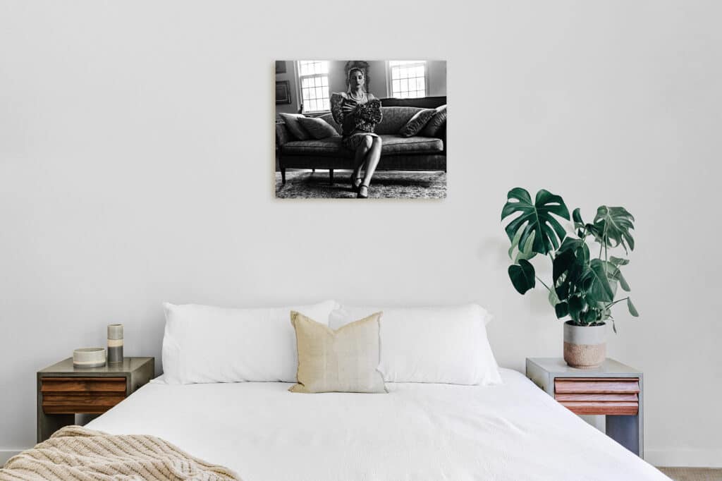 A bright, minimalist bedroom featuring a white bed, a potted Monstera plant on a nightstand, and a black-and-white canvas print of a woman sitting on a sofa hanging on the wall above the bed.