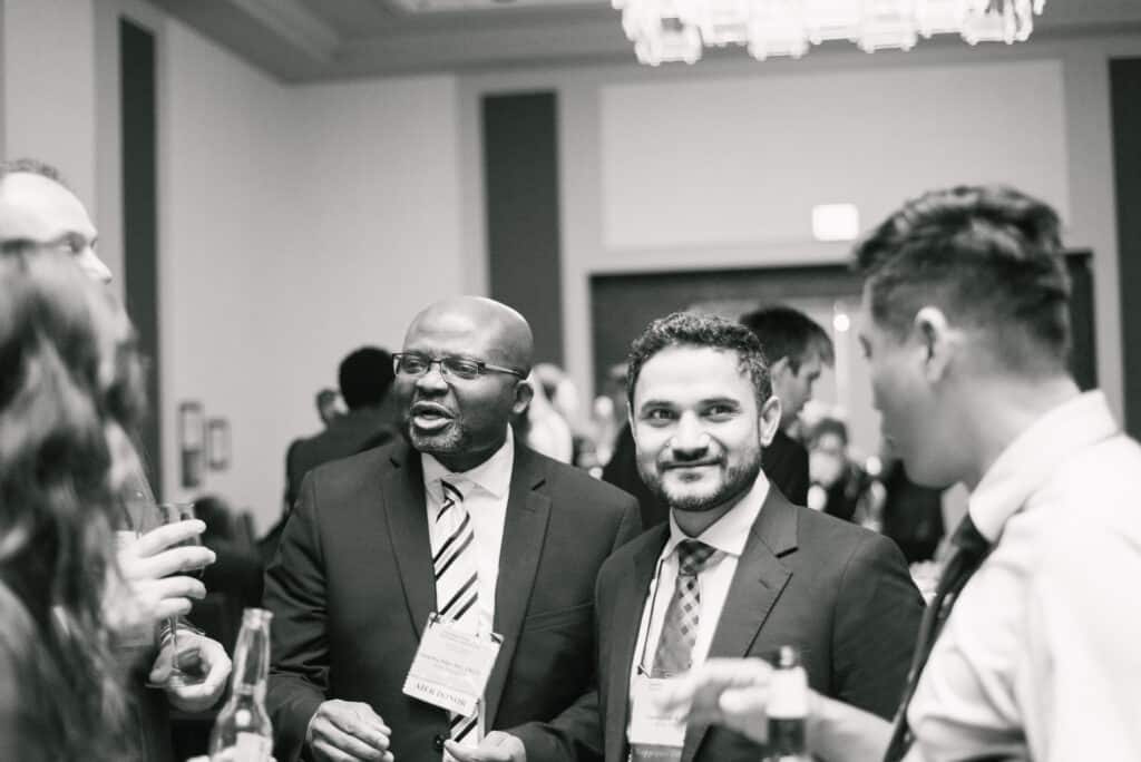 Black and white photo of a group of professionals in suits talking at a networking event, focusing on two men wearing event name badges in the center.