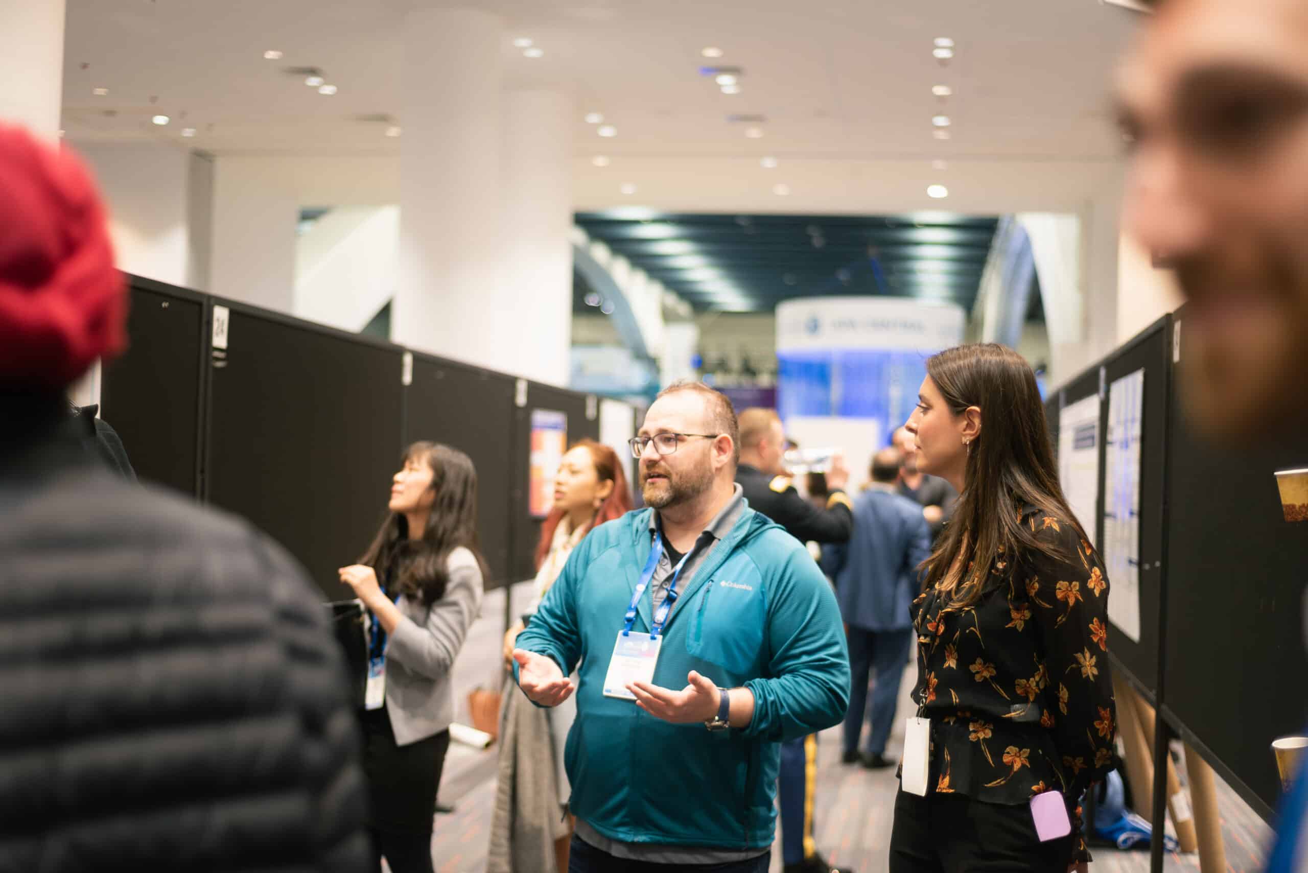 A man in a teal jacket and glasses gestures while speaking to a woman in a floral blouse at a bustling conference poster session, with other attendees and black display boards in the background.