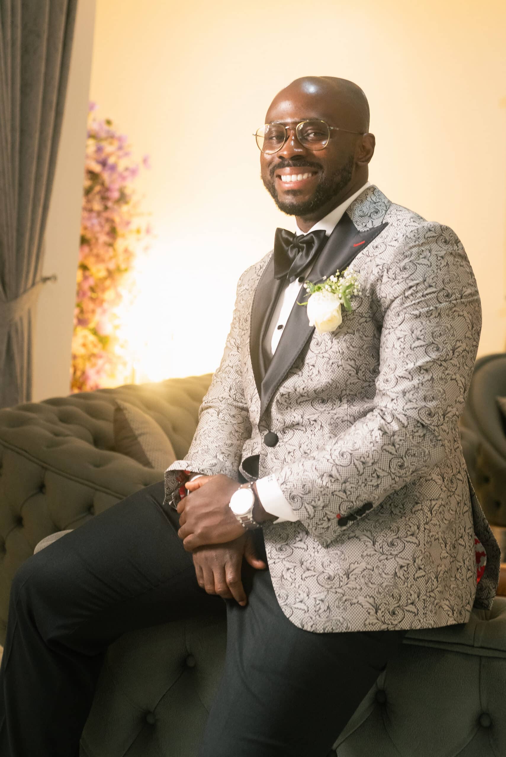 A smiling, sharply dressed groom with glasses wearing an ornate grey patterned tuxedo jacket and black bow tie, perched on the edge of a tufted grey sofa.