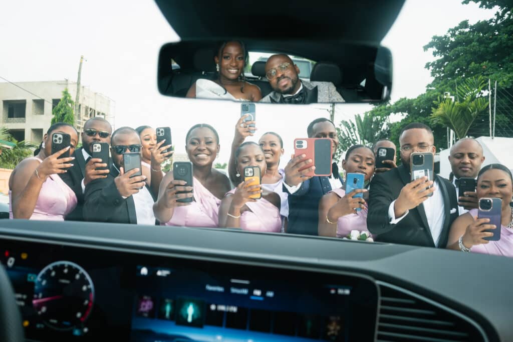 Bridal party taking photos with smartphones of the bride and groom visible in the car's rearview mirror.