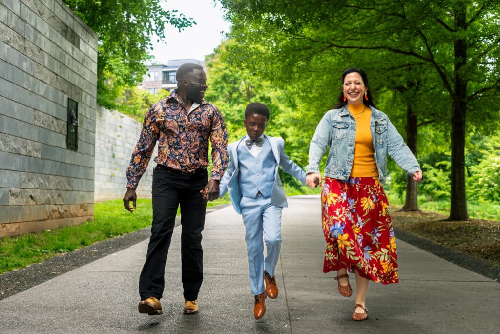 A joyful man, a young boy skipping in a light blue suit, and a smiling woman holding hands as they walk down a tree-lined paved path.
