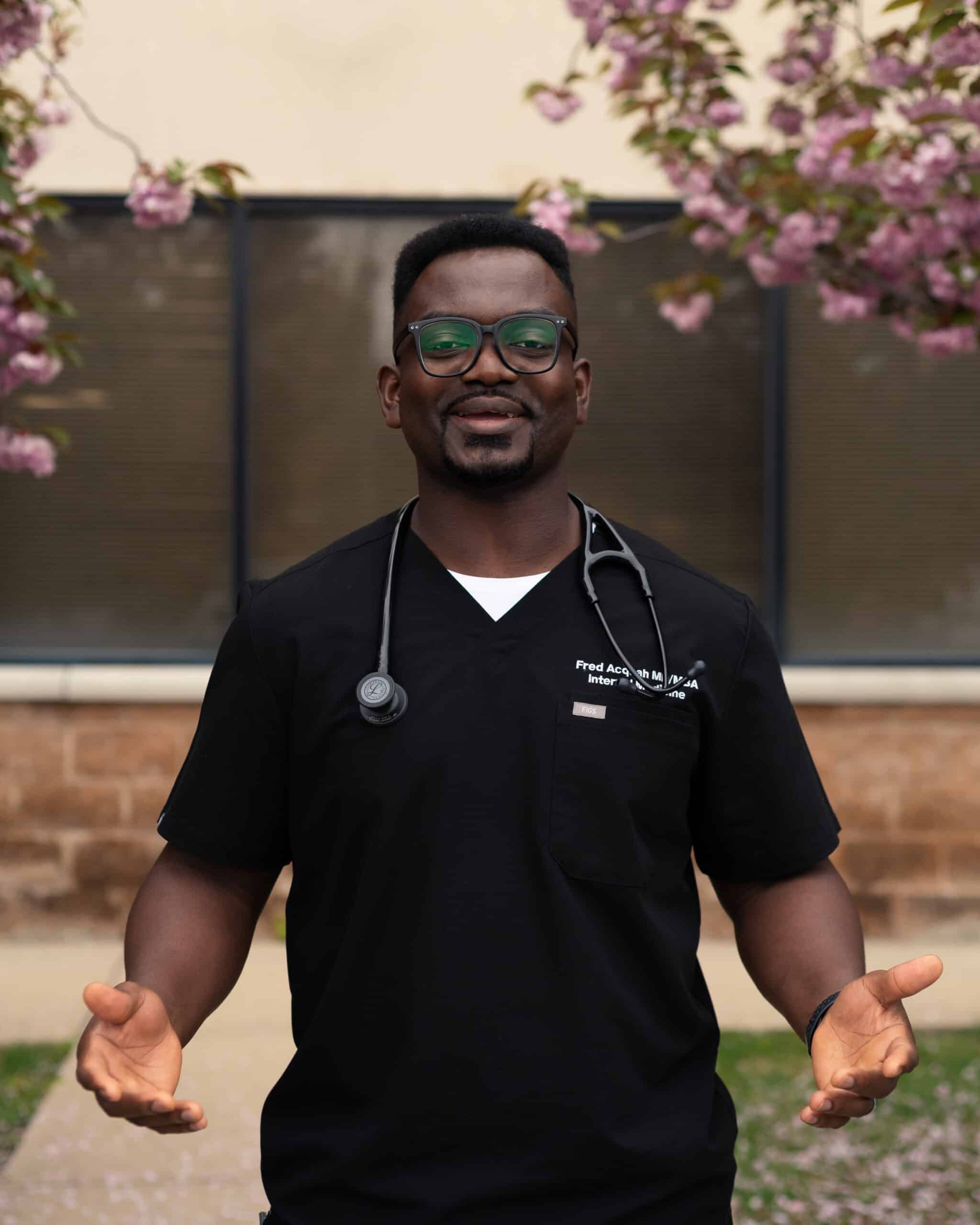 A smiling male doctor wearing black scrubs, glasses, and a stethoscope around his neck, standing outdoors with his hands open in a welcoming gesture, framed by pink spring blossoms.