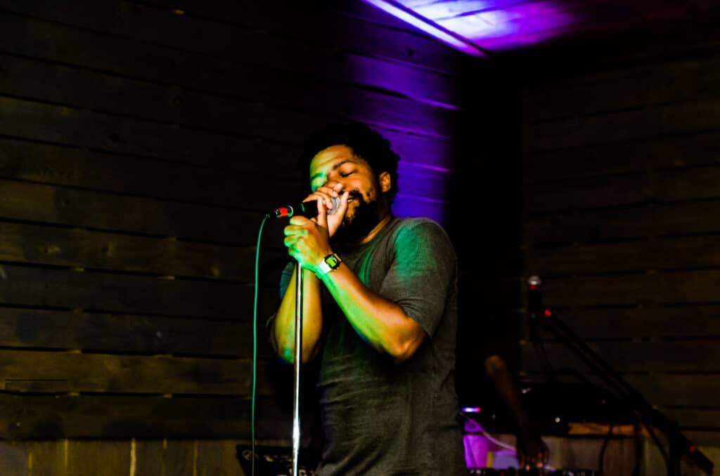 A man with a beard singing passionately into a microphone on stage, illuminated by striking green and purple lights against a dark wooden wall.