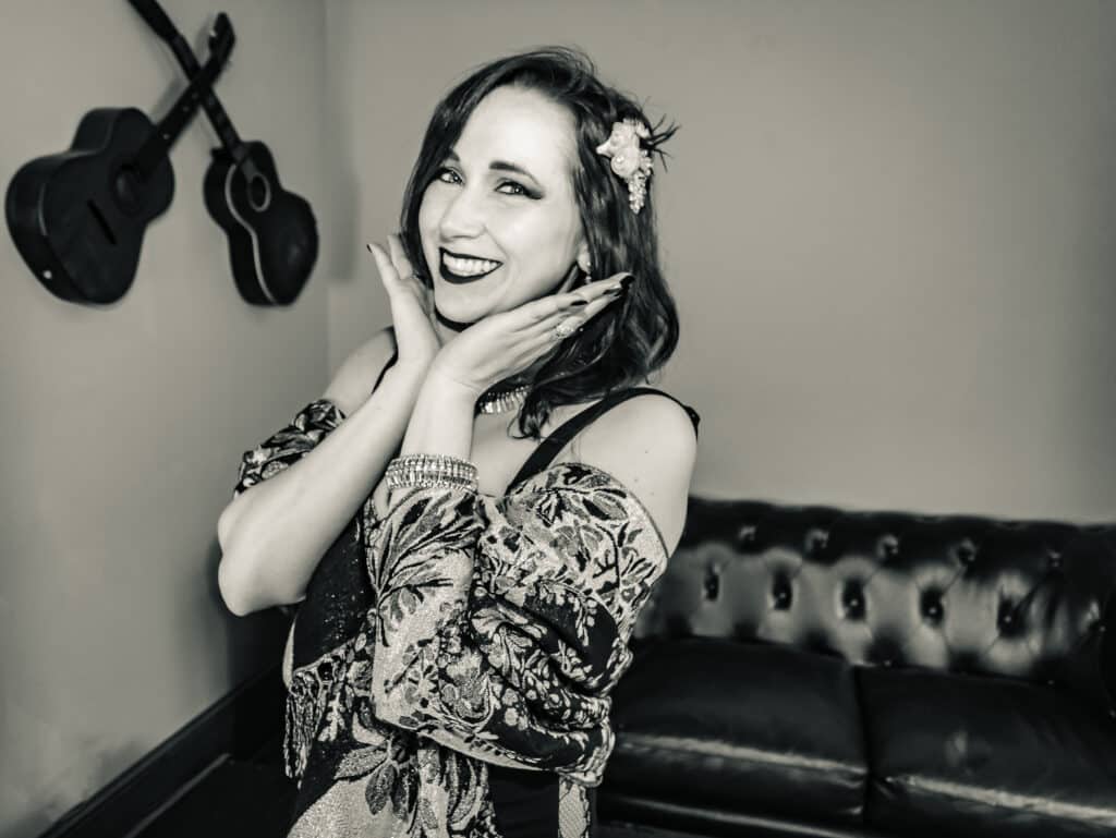 Black and white portrait of a smiling woman wearing a patterned shawl and floral hair accessory, framing her face with her hands.