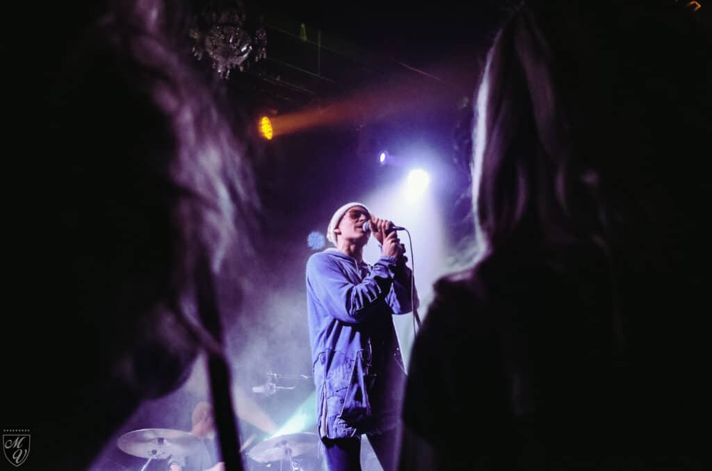 A singer wearing a beanie, sunglasses, and a blue jacket performing on stage, viewed from the audience through the dark silhouettes of crowd members, illuminated by bright purple and white stage lights.