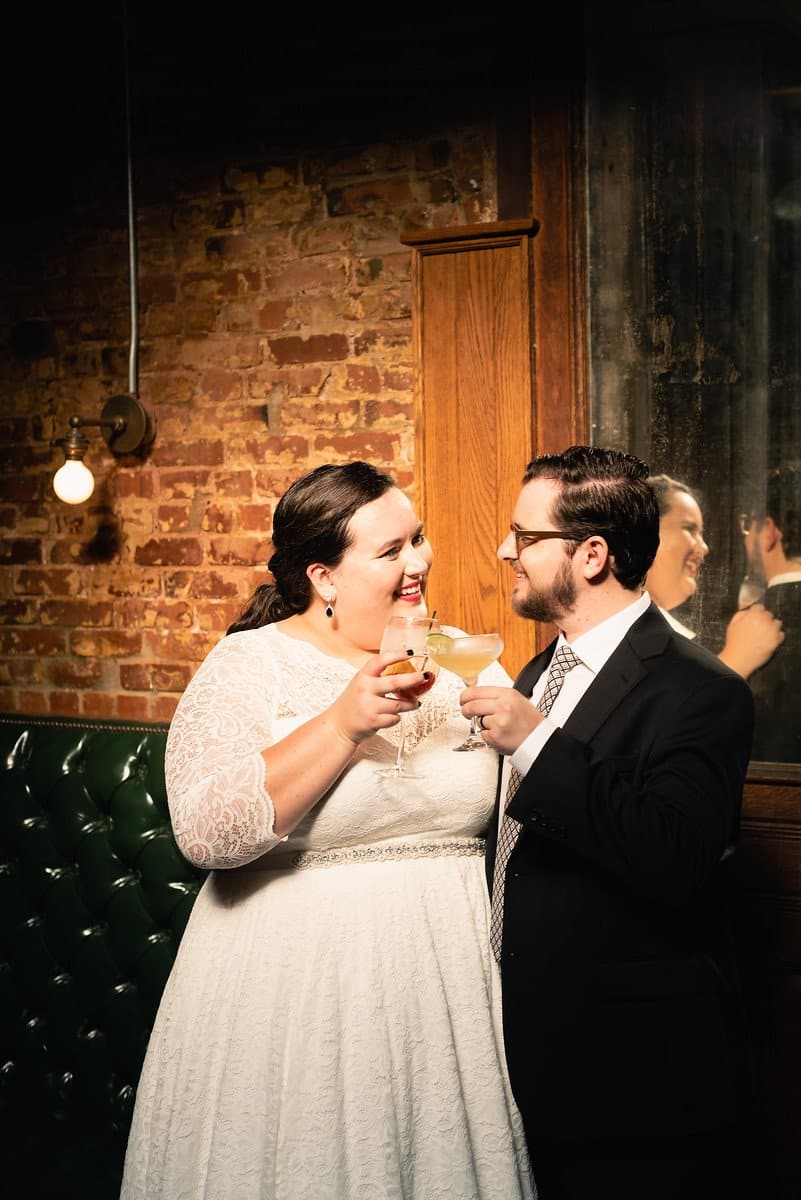 A newlywed couple smiling warmly at each other while clinking their cocktail glasses together inside a dimly lit bar with an exposed brick wall.