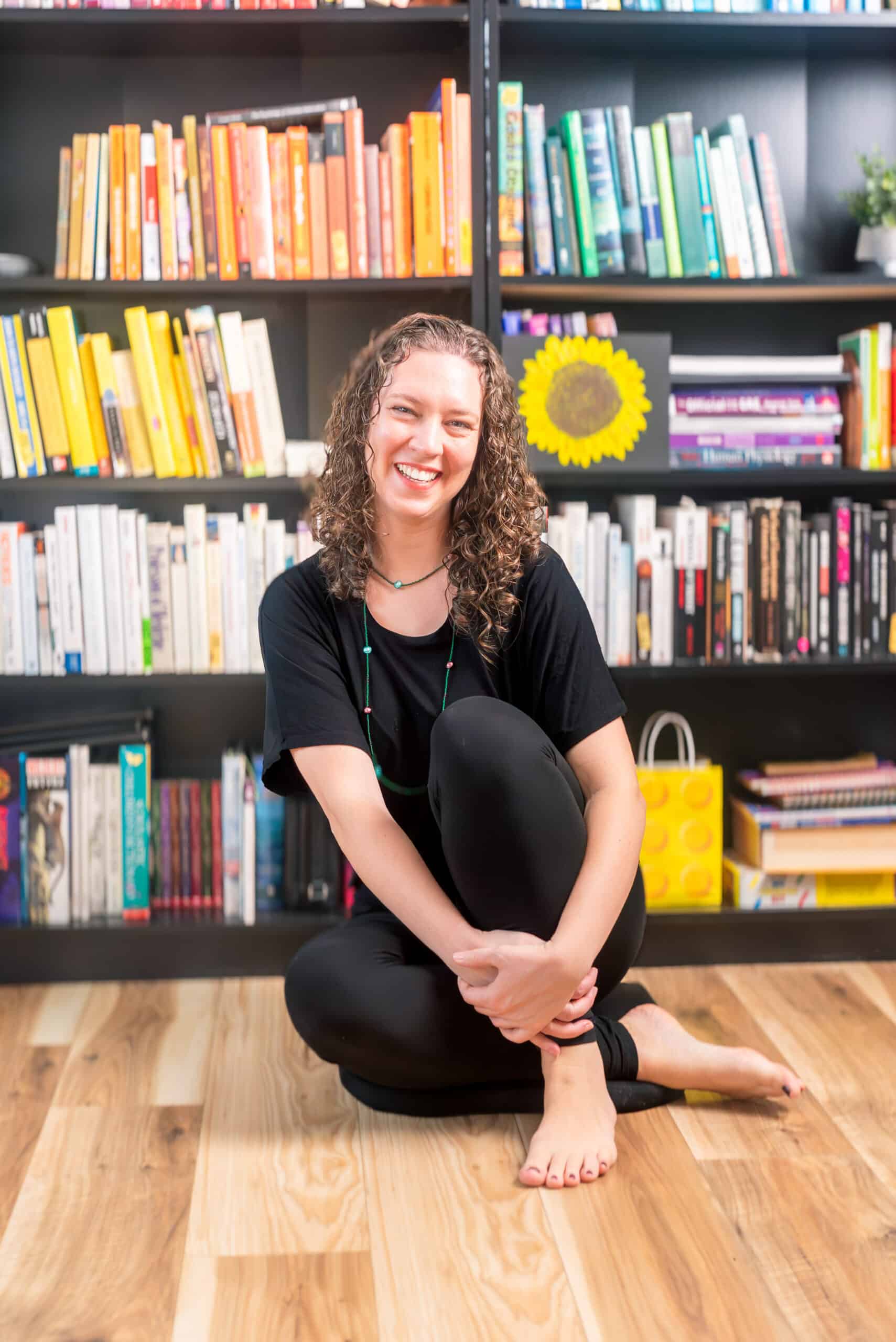 A smiling woman with curly hair sitting barefoot on a wooden floor in front of a black bookshelf filled with color-coordinated books.