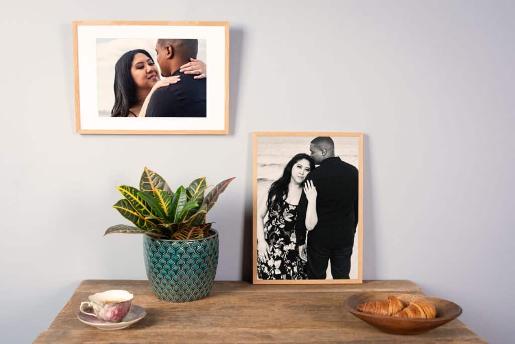 A cozy home decor setup featuring a wooden table with a vintage teacup, a potted croton plant, a bowl of croissants, and two light wood framed photos of a couple.